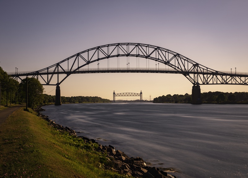 A large steel arch bridge spans a wide waterway at sunset, with a smaller bridge visible in the distance.
